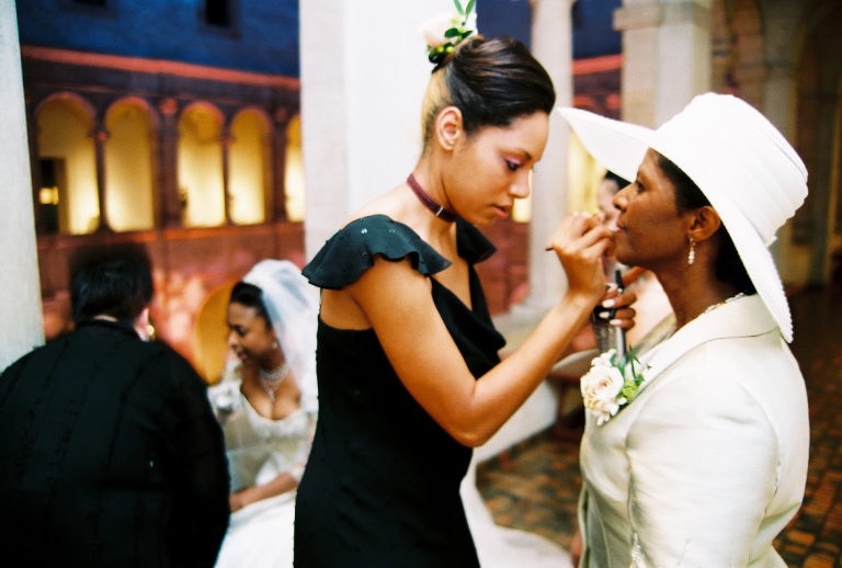 A tall Black woman makeup artist applies lip color to a Black mother of the bride.