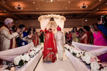 A father walks his daughter up a lavishly decorated wedding aisle to the mandap or wedding stage.