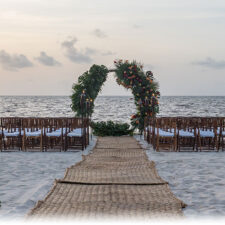 ID: natural fabric carpet runner on the beach, leading to wedding ceremony chairs and ceremony arch setup overlooking the ocean.