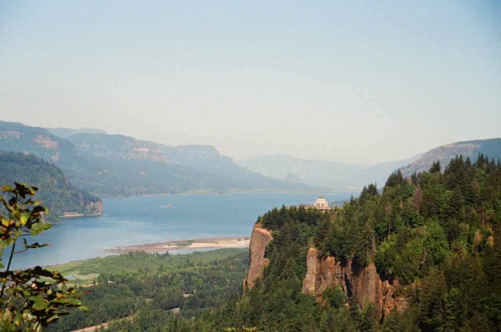 A view of the Columbia River with the forested cliffs in the foreground and the distance, and with a view of the historic Vista House.