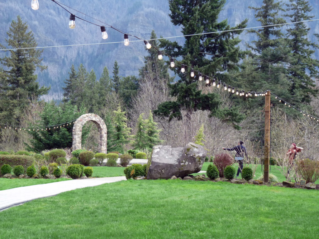 A view of an expansive lawn with a stone arch to mark a ceremony site, decorated with cafe lights above. Two wedding planners talk to each other in the background. An aisle path leads to the arch.