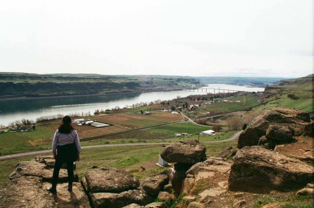 A woman stands on a rocky cliff and looks at the Columbia River and farmlands below